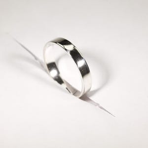 May include: A shiny, silver ring with a flat band. The ring is set against a white background, with a shadow cast to the side. The ring appears to be made of metal and has a polished finish.