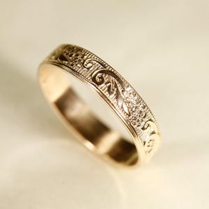 May include: A rose gold band ring featuring an intricate engraved pattern of swirling scrolls and small floral motifs along the outer surface. The metal has a polished finish, highlighting the detailed texture of the embossed design across the band.