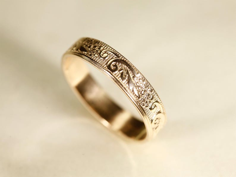 May include: A rose gold band ring featuring an intricate engraved pattern of swirling scrolls and small floral motifs along the outer surface. The metal has a polished finish, highlighting the detailed texture of the embossed design across the band.