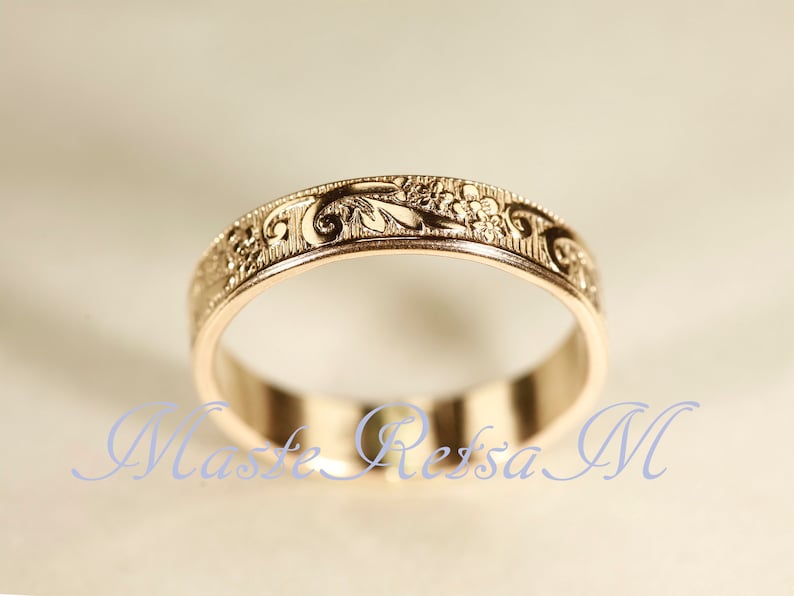 May include: A gold band ring featuring an intricate engraved floral pattern along the outer surface. The metal has a polished finish with detailed scrollwork and small flower motifs etched into the metal. The text Maste Retsa M is visible.