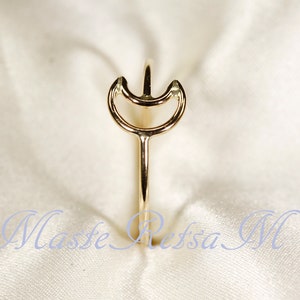 May include: A gold-colored ring with a unique design. The ring features a slender band that extends upwards, culminating in a stylized crescent shape with a central point. The ring is set against a soft, white background.