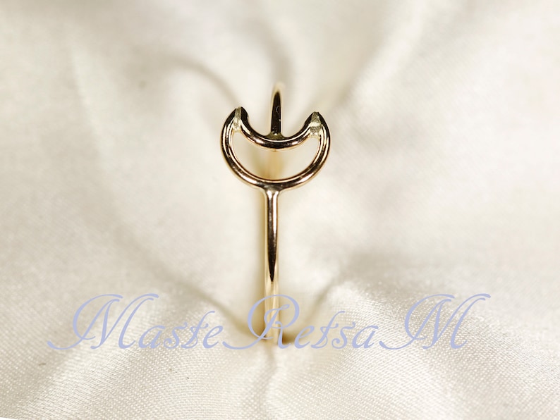 May include: A gold-colored ring with a unique design. The ring features a slender band that extends upwards, culminating in a stylized crescent shape with a central point. The ring is set against a soft, white background.