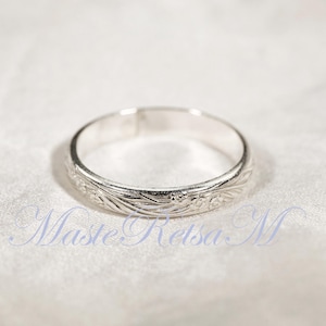 May include: A silver ring with a floral pattern. The ring is circular and has a detailed design of leaves and small flowers. The ring is displayed on a white, textured surface.