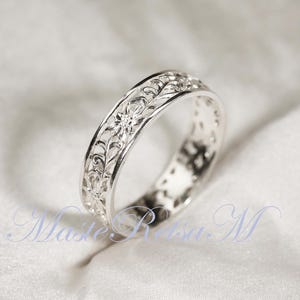 May include: A silver ring with an intricate floral filigree design. The ring's band features delicate cut-out patterns and detailed floral motifs. The ring is displayed on a white, textured surface, highlighting its reflective qualities.