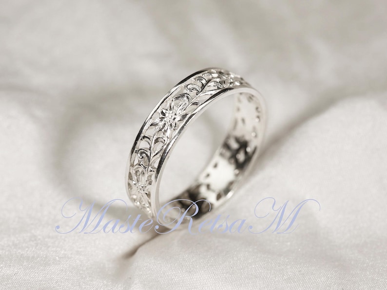 May include: A silver ring with an intricate floral filigree design. The ring's band features delicate cut-out patterns and detailed floral motifs. The ring is displayed on a white, textured surface, highlighting its reflective qualities.