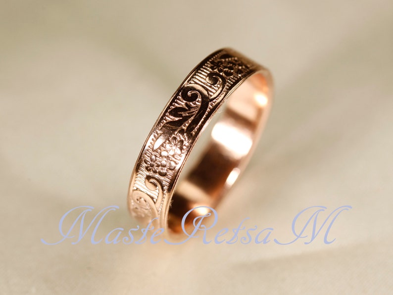 May include: A rose gold ring featuring an engraved floral pattern with swirling vines and small blossoms. The band has a polished finish and a flat profile, showcasing intricate metalwork that wraps around the entire circumference of the ring.