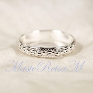 May include: A silver ring with an intricate, repeating pattern around the band. The ring is set against a soft, white background. The ring's design includes a series of diamond and dot shapes. The text "Maste Retsa M" is visible in the image.