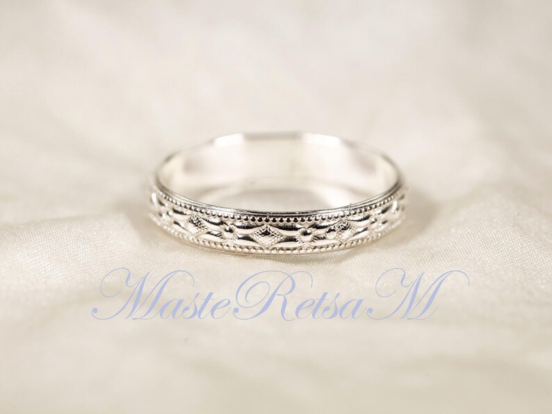 May include: A silver ring with an intricate, repeating pattern around the band. The ring is set against a soft, white background. The ring's design includes a series of diamond and dot shapes. The text "Maste Retsa M" is visible in the image.