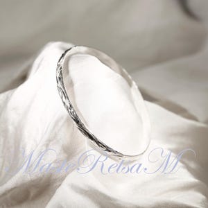 May include: A silver bangle bracelet with a textured, twisted design. The bracelet is on a white fabric background.