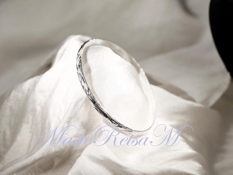 May include: A silver bangle bracelet with a textured, twisted design. The bracelet is on a white fabric background.