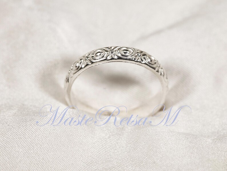 Puede incluir: Un anillo plateado con un intrincado dise&ntilde;o floral. La superficie del anillo est&aacute; adornada con un patr&oacute;n repetitivo de espirales y motivos florales. El anillo est&aacute; sobre un fondo blanco, resaltando el dise&ntilde;o detallado.