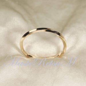 Może przedstawiać: Złoty pierścionek ze skręconym wzorem. Pierścionek jest umieszczony na miękkim, kremowym tle. Obręcz pierścionka ma gładkie, wypolerowane wykończenie, odbijające światło. Pierścionek to prosty, elegancki element biżuterii.