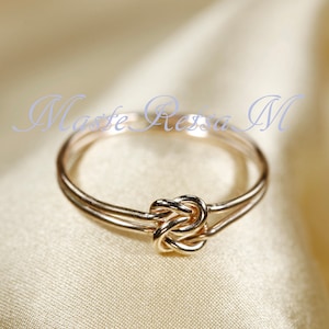 May include: A delicate gold-colored ring with a knot design at the center. The ring has a double band and a polished finish. The background is a soft, neutral color, highlighting the ring's intricate details. The text "Masse Ressa M" is visible in the background.