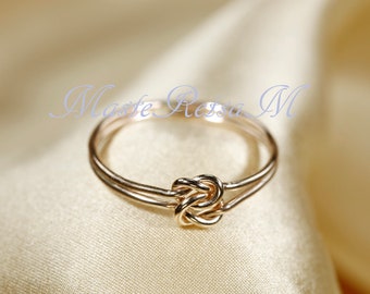 May include: A gold-coloured ring featuring a knot design at the centre. The ring has a double band and a polished finish. The background is a soft, neutral colour, highlighting the ring's intricate details. The text "Masse Ressa M" is visible in the background.
