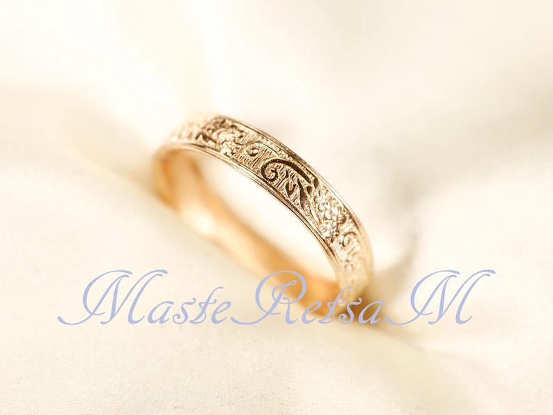 May include: A gold ring featuring an intricate, embossed floral and scrollwork pattern along its outer band. The metal has a warm, polished finish, and the design consists of repeating organic shapes and fine lines engraved across the surface.