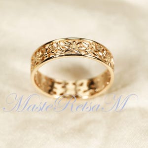 May include: A gold-colored ring with an intricate floral design. The ring features a continuous pattern of flowers and leaves around the band. The ring is set against a soft, neutral background. The ring is a piece of jewelry.