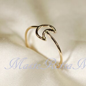 May include: A delicate gold-colored ring with a crescent moon design. The ring features a thin band that curves into a stylized moon shape. The ring is set against a soft, off-white background. The text "Maste Ressa M" is visible in the image.