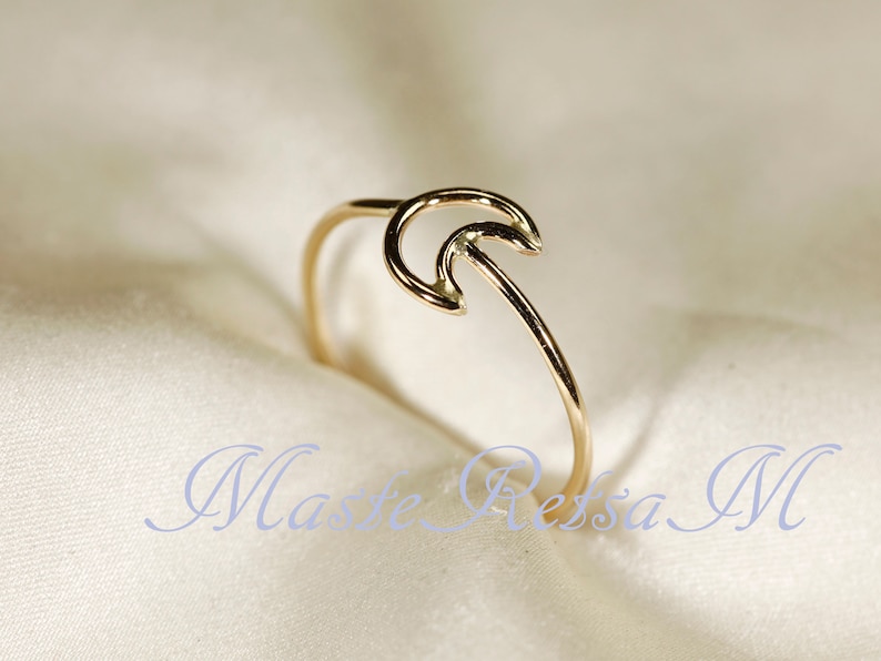 May include: A delicate gold-colored ring with a crescent moon design. The ring features a thin band that curves into a stylized moon shape. The ring is set against a soft, off-white background. The text "Maste Ressa M" is visible in the image.