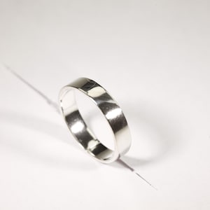 Puede incluir: Un anillo de plata con una banda plana, sobre una superficie blanca. El anillo refleja la luz, creando reflejos y sombras. La joya es simple y elegante, adecuada para el uso diario. El diseño del anillo es minimalista.