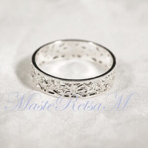 May include: A silver ring with a floral filigree design. The ring has an openwork pattern of flowers and leaves around the band. The ring is on a white surface.