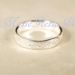 May include: A silver ring featuring an intricate, engraved floral pattern along its outer band. The metal has a polished, reflective finish. The text Maste Retsa M is superimposed in light blue script across the upper portion of the image.