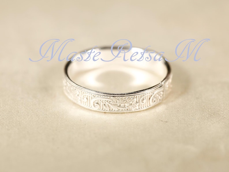 May include: A silver ring featuring an intricate, engraved floral pattern along its outer band. The metal has a polished, reflective finish. The text Maste Retsa M is superimposed in light blue script across the upper portion of the image.