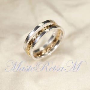 Peut inclure: Une bague en argent avec une bande centrale dorée et un motif texturé. La bague a une finition polie et est posée sur un fond doux et neutre. Il s'agit d'un bijou.