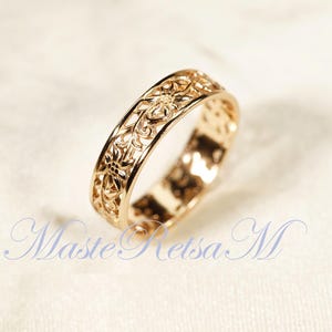 May include: A gold-colored ring with an intricate floral filigree design. The ring is made of a shiny, polished metal and features a continuous pattern of flowers and leaves. The ring is shown on a white surface.