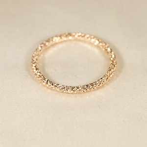 May include: A thin, circular gold ring featuring a highly textured, faceted surface that creates a sparkling, glitter-like appearance across the entire band. The metal has a warm, metallic yellow tone with numerous small, reflective indentations.