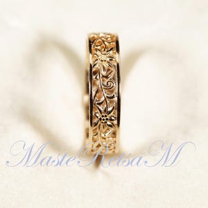 May include: A gold-colored ring with an intricate floral design. The ring features a continuous pattern of flowers and vines, creating a delicate, openwork effect. The ring is likely made of gold and is a piece of jewelry.