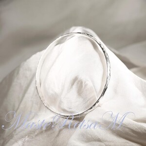 May include: A silver bangle bracelet with a hammered texture. The bracelet is lying on a white fabric surface.