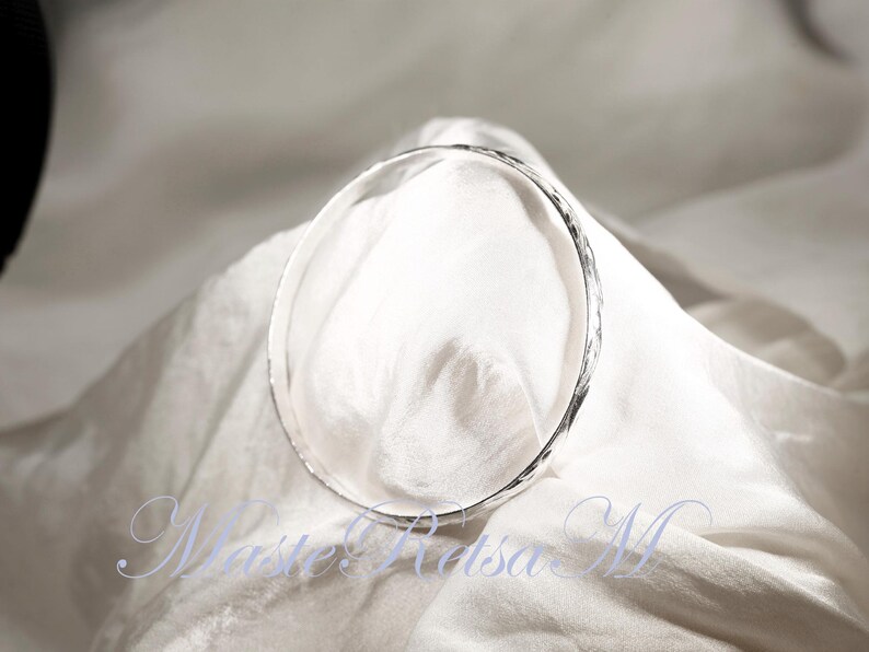 May include: A silver bangle bracelet with a hammered texture. The bracelet is lying on a white fabric surface.