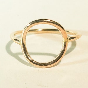 May include: A gold-toned ring featuring a thin, smooth band and a prominent circular centerpiece. The metal has a polished, reflective finish that catches the light, creating a simple, continuous loop design that sits centered on the band.