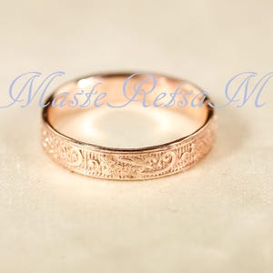 May include: A rose gold ring featuring an intricate, embossed floral scrollwork pattern around the entire outer band. The text Maste Retsa M is overlaid in blue script across the center of the ring, which has a polished, metallic finish.