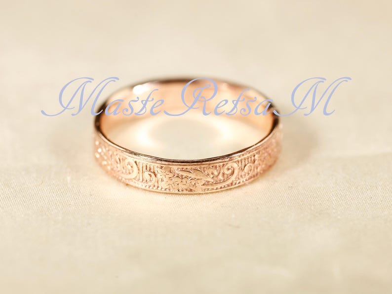 May include: A rose gold ring featuring an intricate, embossed floral scrollwork pattern around the entire outer band. The text Maste Retsa M is overlaid in blue script across the center of the ring, which has a polished, metallic finish.