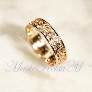 May include: A gold-colored ring with an intricate floral filigree design. The ring's band is adorned with detailed cut-out patterns of flowers and leaves. The ring is displayed on a soft, light-colored surface. The text "Maste Retsa M" is visible.