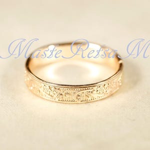 May include: A rose gold ring featuring an intricate engraved floral pattern along the outer band. The text "Maste Retsa M" is overlaid in blue script across the top of the ring. The metal has a polished finish that reflects light.