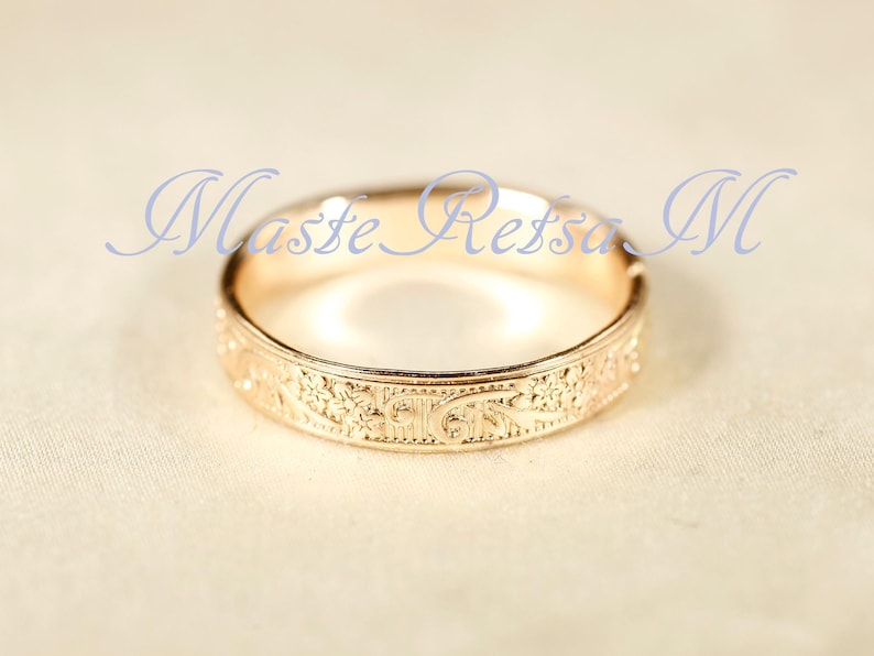 May include: A rose gold ring featuring an intricate engraved floral pattern along the outer band. The text "Maste Retsa M" is overlaid in blue script across the top of the ring. The metal has a polished finish that reflects light.
