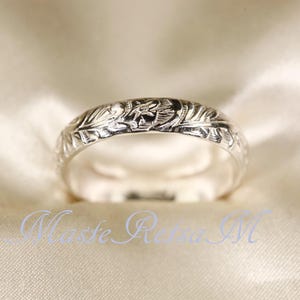 Può includere: Un anello d'argento con un motivo floreale inciso. L'anello ha una finitura lucida ed è posto su uno sfondo bianco sporco. Il design dell'anello include foglie e un elemento floreale centrale.