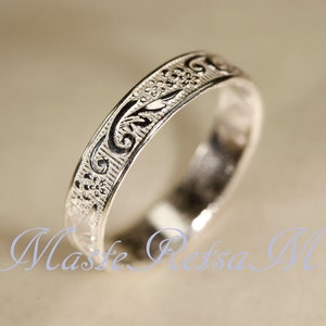 May include: A silver-toned metal ring featuring an intricate embossed pattern of small flowers and swirling vine-like motifs across its outer surface. The design includes dark recessed areas that highlight the raised floral and scrollwork details.