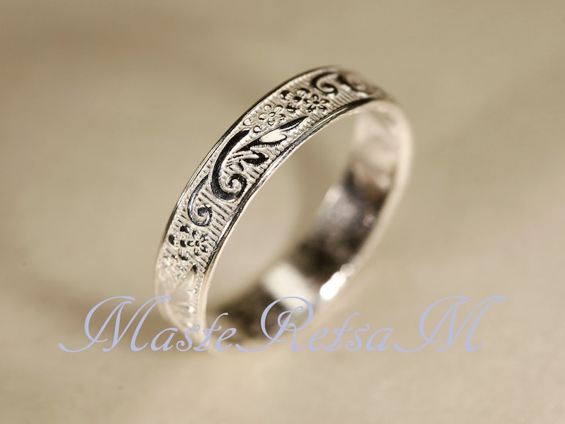 May include: A silver-toned metal ring featuring an intricate embossed pattern of small flowers and swirling vine-like motifs across its outer surface. The design includes dark recessed areas that highlight the raised floral and scrollwork details.
