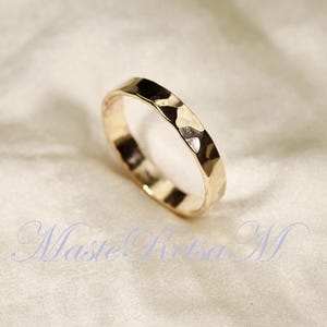 May include: A gold-colored ring with a hammered texture. The ring has a faceted surface that reflects light, creating a sparkling effect. The ring is set against a soft, off-white background.