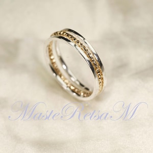 Könnte beinhalten: Ein silberner Ring mit einem goldenen Band und einem strukturierten goldenen Mittelband. Der Ring hat ein Doppelband-Design und ist vor einem weichen, cremefarbenen Hintergrund platziert. Der Ring ist ein Schmuckstück.