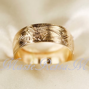 May include: A gold ring featuring an embossed floral pattern with swirling vine details across the outer band. The surface of the metal has a textured finish. The text "Masse Retsa M" is overlaid in light blue script across the lower portion.