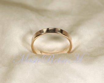 May include: A gold-colored band ring with a flat, polished surface. The ring is set against a soft, off-white fabric, creating a simple and elegant presentation. The ring's design is minimalist and classic.