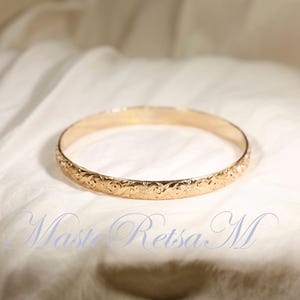 Puede incluir: Un brazalete de color dorado con un patrón floral grabado. El brazalete es circular y descansa sobre una superficie blanca y texturizada. El diseño del brazalete es visible alrededor de toda la circunferencia.