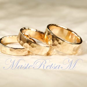 May include: Three gold-colored rings with a hammered texture are arranged on a light-colored surface. The rings are in focus, with a soft background. The rings have a textured, hammered finish. The rings are likely wedding bands or fashion rings.