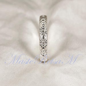 Puede incluir: Un anillo plateado con un intrincado patr&oacute;n repetitivo de dise&ntilde;os ovalados y en forma de lazo. El anillo se presenta sobre un fondo blanco suave, resaltando su detallada artesan&iacute;a. El dise&ntilde;o del anillo es sim&eacute;trico y elegante.