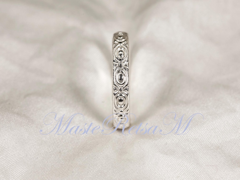 Puede incluir: Un anillo plateado con un intrincado patr&oacute;n repetitivo de dise&ntilde;os ovalados y en forma de lazo. El anillo se presenta sobre un fondo blanco suave, resaltando su detallada artesan&iacute;a. El dise&ntilde;o del anillo es sim&eacute;trico y elegante.