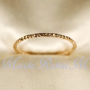 Könnte beinhalten: Ein goldfarbener Ring mit strukturierter Oberfläche. Der Ring hat ein sich wiederholendes geometrisches Muster um das Band. Der Ring wird auf einem cremefarbenen Stoff präsentiert.